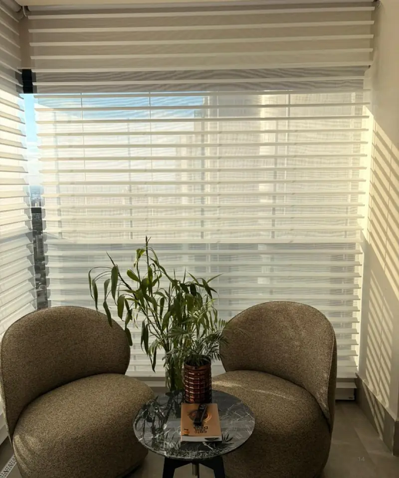 Cortina de lâminas de tecido em ondas, translúcida, em uma sala com poltronas e planta.