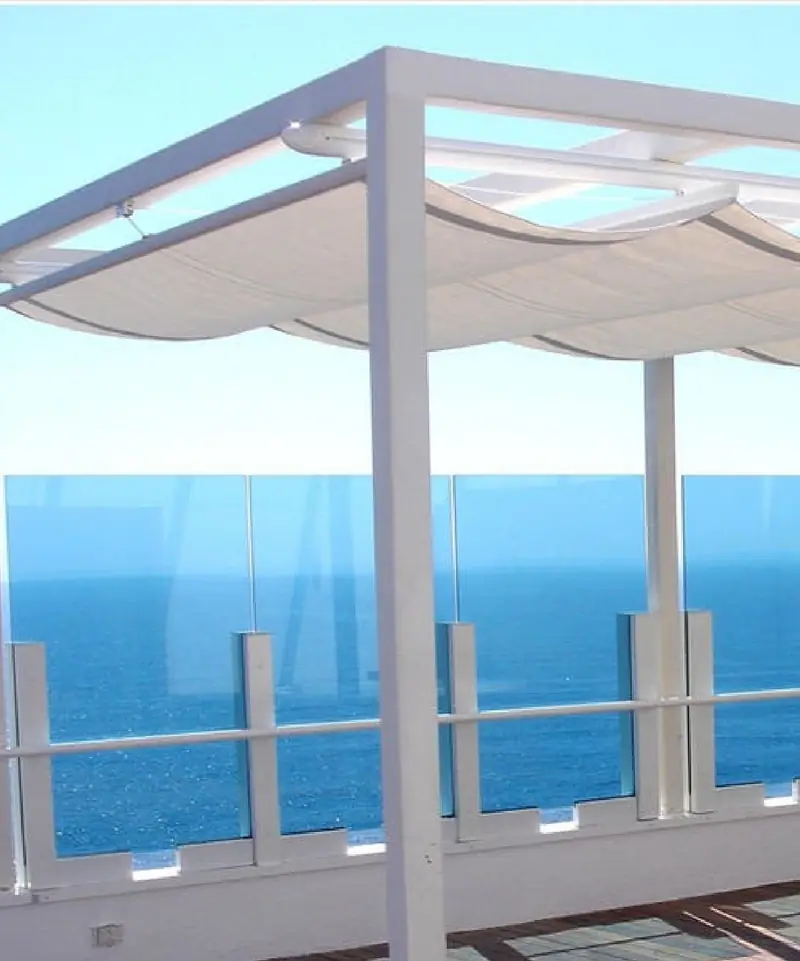 Toldo sobre pergolado em uma varanda com guarda-corpo de vidro e vista para o mar.