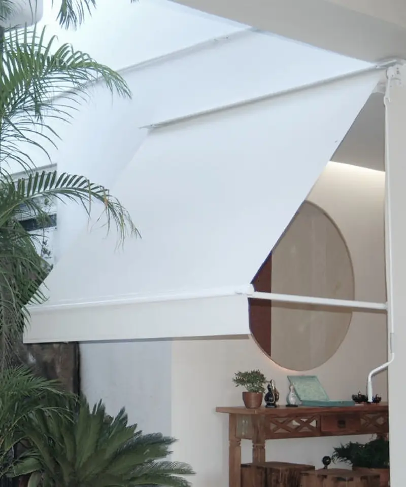 Toldo branco com braços pivotantes aberto em um ambiente com plantas ao fundo.